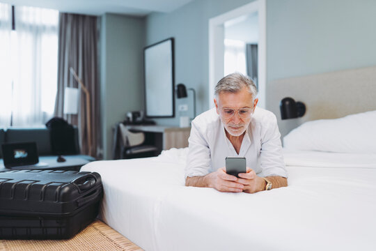 Man with eyeglasses lying on bed while using smart phone in hotel room