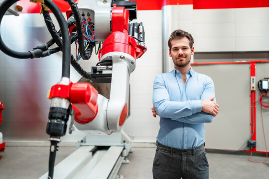 Male engineer with arms crossed standing at robotics in factory