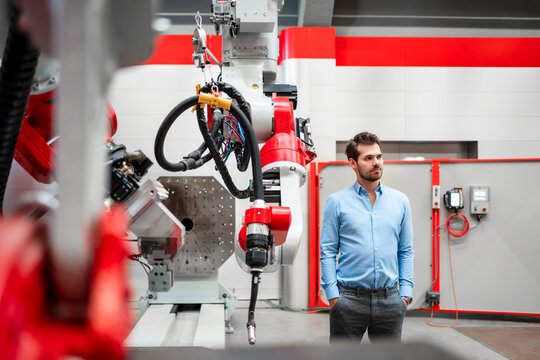 Male engineer with hands in pockets looking away while standing at robotics in factory