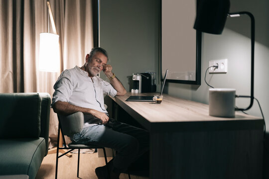 Tired man rubbing eyes while sitting at desk against laptop in hotel room