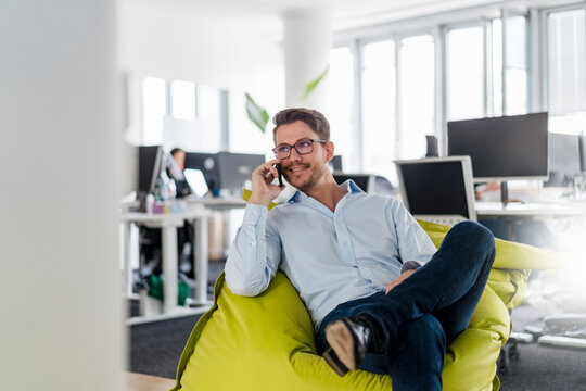 Handsome businessman talking through smart phone while sitting on bean bag at office - Powered by Adobe