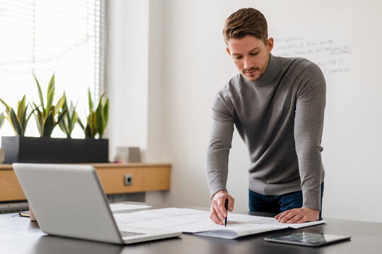 Male entrepreneur analyzing blueprint by laptop at desk in office