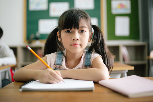 Portrait of little Asian girl writing or drawing in notebook at desk in classroom and looking at camera at the elementary school. Education knowledge, activities with kids concept.