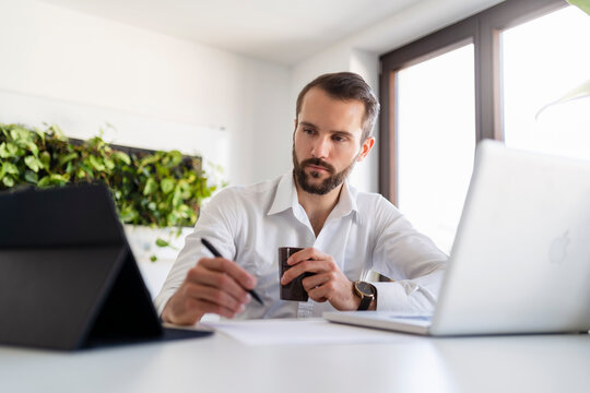 Businessman with laptop and coffee cup using digital tablet at office - Powered by Adobe