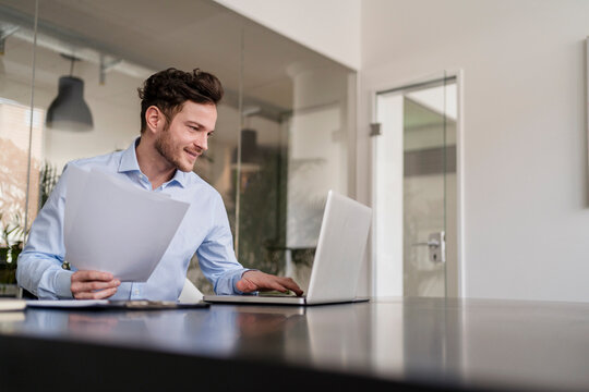 Smiling male entrepreneur with document working on laptop in office