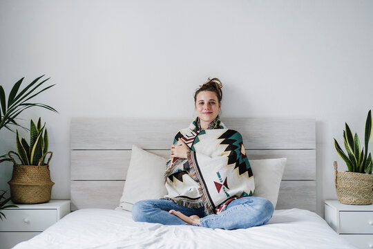Young woman wrapped in shawl smiling while sitting on bed at home