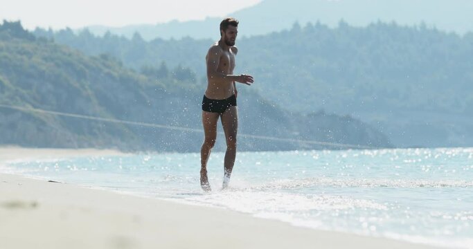 The handsome man with a perfect athletic body in swimming trunks having fun on a deserted beach in the morning, splashing in the water, playing with the waves, showing off his body and posing