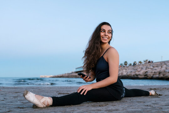 Smiling ballet dancer looking over shoulder while doing leg split at beach against clear sky