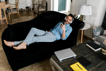 Smiling man with headphones using smart phone while relaxing on sofa at home