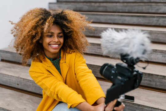 Smiling Afro woman holding camera during vlogging while sitting on staircase