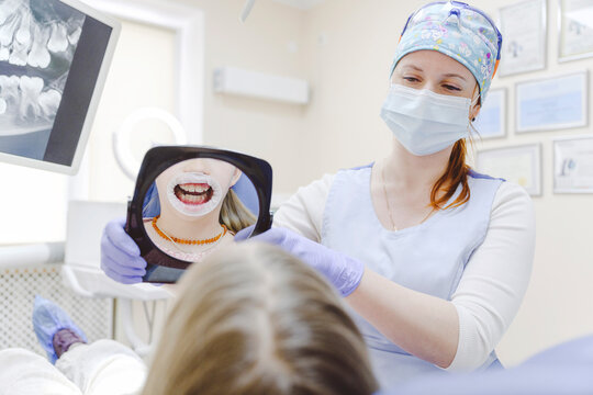 Female dentists holding mirror reflecting mouth of little girl wearing dental gag