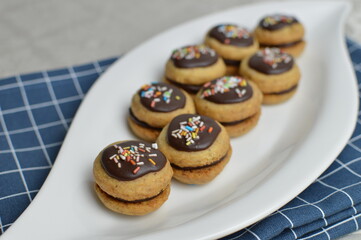 Mini traditional Austrian sandwich cookies Ischler (isleri) with chocolate and colorful sprinkles on the oval white plate and blue kitchen cloth 