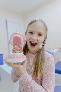 Portrait of little blonde girl standing in dentists office with dentures in hands