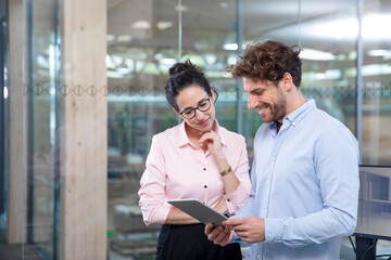 Young male entrepreneur with digital tablet discussing with female colleague against glass wall at office