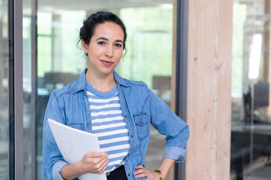 Confident female entrepreneur with hand on hip holding document while leaning on glass wall at office