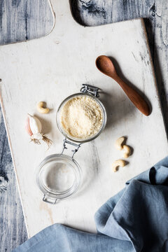 Cutting board with napkin, wooden spoon, garlic, cashews and jar of shredded vegan Parmesan cheese