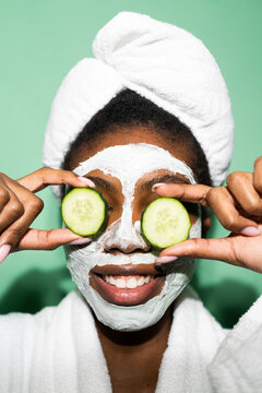 Smiling woman with facial mask covering eyes with cucumber while standing against green background