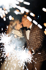 groom in a jacket and a bride in a jacket are holding burning sparklers and fireworks in their hands. Kisses against the background of sparks and splashes of flashes.