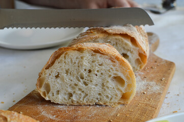 Slicing homemade sourdough rustic artisan baguette with bread knife on the wooden bread board, side view