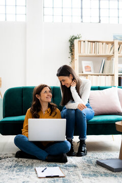 Mother with laptop smiling at daughter while sitting at sofa in living room