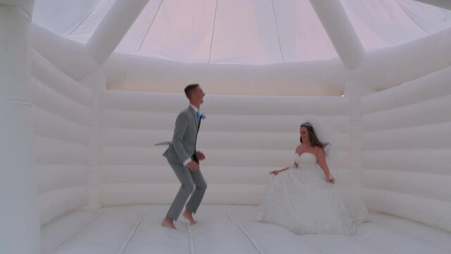 Whimsical moment as a young bride and groom playfully jump around in a white bouncy house while still wearing their wedding dress and tuxedo.