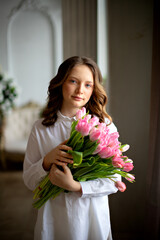 A beautiful little girl in a white shirt with long curly brown hair and blue eyes holds a huge bouquet of Tulips in her hands