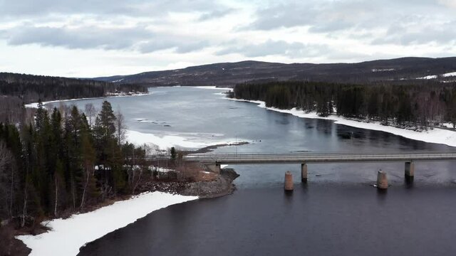 River in swedish winter. Bridge with car crossing. Indalsälven water stream. Forest and now in nature. Cloudy evening in Sweden nature. flying aerial drone shot. Åre Undersåker jämtland