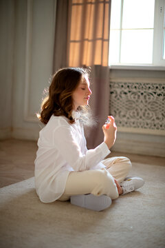 A Beautiful Little Girl In A White Shirt With Long Curly Brown Hair And Blue Eyes Sits On The Floor And Sprays Perfume On Herself