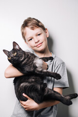 Boy with blond hair and in a gray t-shirt hugs a cute dark cat on a white background