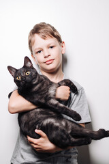 Boy with blond hair and in a gray t-shirt hugs a cute dark cat on a white background