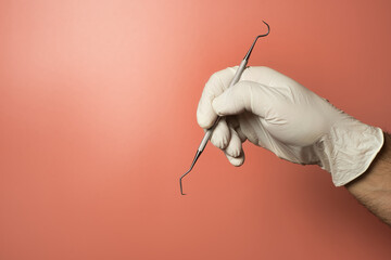 Faceless dentist wearing latex gloves holding periodontal scaler against a light red background.
