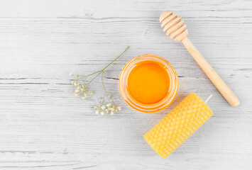 Beekeeping products. Open jar with honey, beeswax candle, wooden  honey dipper and a flower on a white wooden background. Top view, copy space. Alternative medicine, vegetarianism, healthy eating.