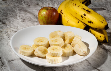 A bunch of bananas, a sliced banana, and an apple on a plate on a table. Selective focus