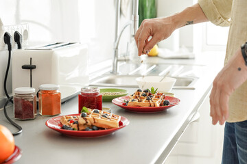 Obraz na płótnie Canvas Male hand of mature man scattering nuts over cooked baked homemade waffles on two plates preparing morning breakfast for couple at home. Close up view