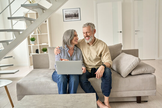 Happy senior mature family couple using laptop computer at home. Smiling mid aged 50s husband embracing wife enjoying online shopping, ordering delivery, buying insurance browsing sitting on sofa.