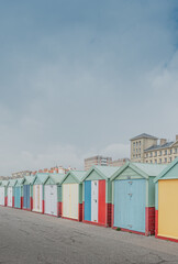 Coloridas casetas de playa en tonos pastel y motivos de rayas, frente a la playa de Brighton, Inglaterra. De fondo unos edificios de estilo clásico en colores claros y un cielo nublado