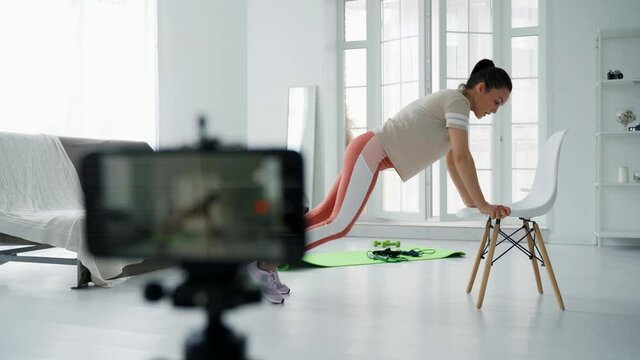 The girl shoots her fitness video blog on a smartphone. The girl conducts distance fitness training online. The girl shoots a video blog during fitness. Physical exercises.