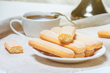 Traditional italian savoiardi biscuits or ladyfingers cookies on a plate and white cup of coffee on background. Selective focus