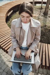 Young beautiful woman working on a laptop sitting on the bench in the street. Freelance business concept.