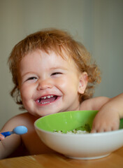 Funny baby eating food himself with a spoon on kitchen. Smiling baby eating food.