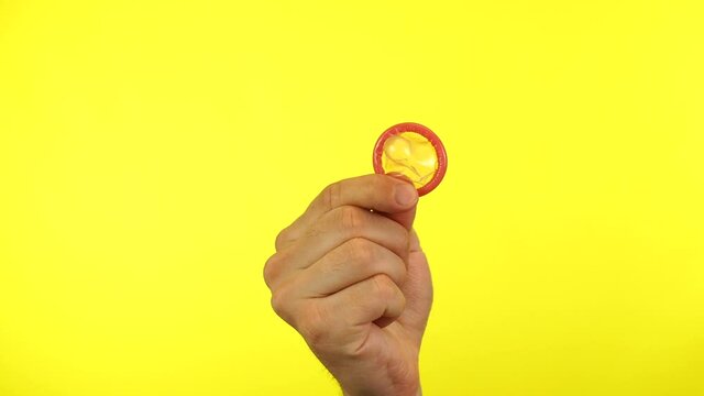 Male hand showing unpacked condom on yellow background. Close up footage of hand only.