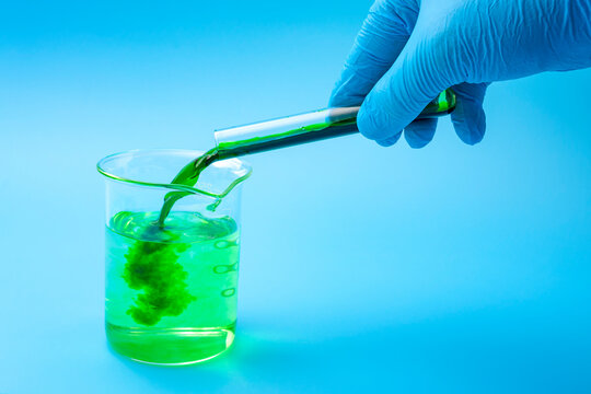 Chemistry, scientific experiment, chemical reaction and mixing chemicals concept with scientist pouring green chemical from test tube in transparent lab flask filled with water on blue background
