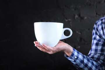 A large white mug in the man's hand. An unrecognizable person. Checkered shirt. A copy of the space.