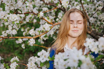 A young woman rejoices in the sun and puts her face to the sun. White apple trees are blooming all around. The blonde has a smile on her face. High quality photo