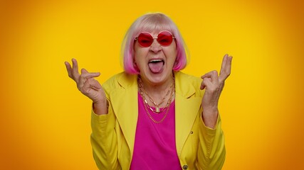Senior old stylish granny woman in sunglasses making playful silly facial expressions and grimacing, fooling around, showing tongue. Mature grandmother isolated on orange background. People lifestyle