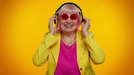 Elderly old granny stylish woman listening music via headphones and dancing disco fooling around, expressive gesticulating hands, having fun. Senior grandmother pensioner on orange studio background