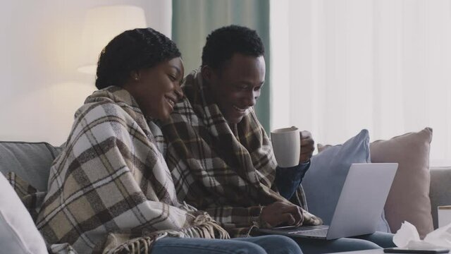 Young sick african american couple wrapped in plaids sitting on sofa together and watching movie on laptop