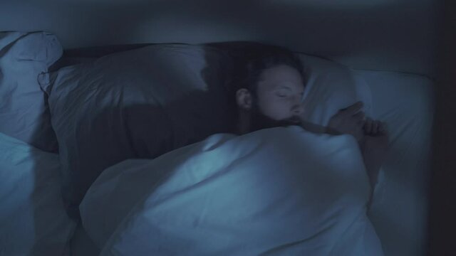 Sleep disorder. Night insomnia. Stress fatigue. Tired bored annoyed man tossing turning covering face with blanket lying in bed awake in dark bedroom with blue moonlight.