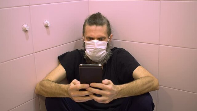 Middle aged bearded man in medical mask shocked bad news of dangerous disease infection watching on smartphone hiding from everyone in bathroom