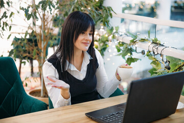 Young beautiful girl communicating on Skype on a laptop computer while sitting in a cafe holding a...